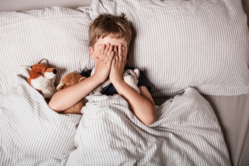 Child covering face while laying in bed with their stuffed animal.