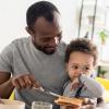 Father with young son on his lap spreading peanut butter on toast.