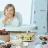 Toddler sitting in a highchair looking at an assortment of cakes and sweets on a table.