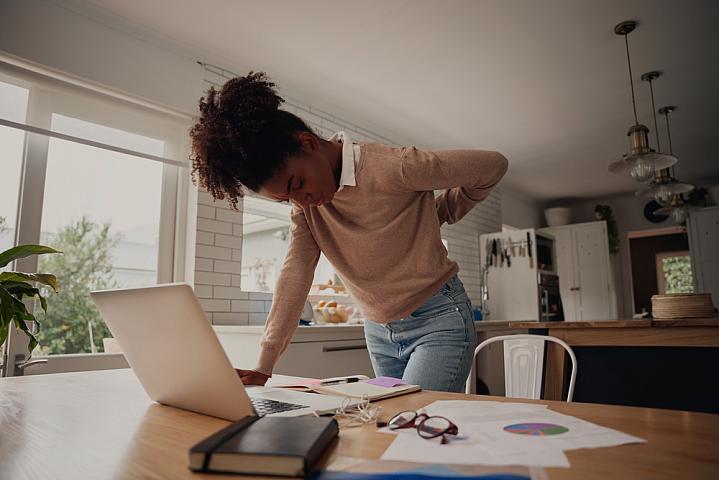 Young woman standing and holding her back while working on a laptop at home