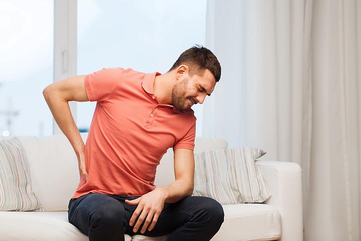 Man sitting on couch holding lower back while wincing in pain.